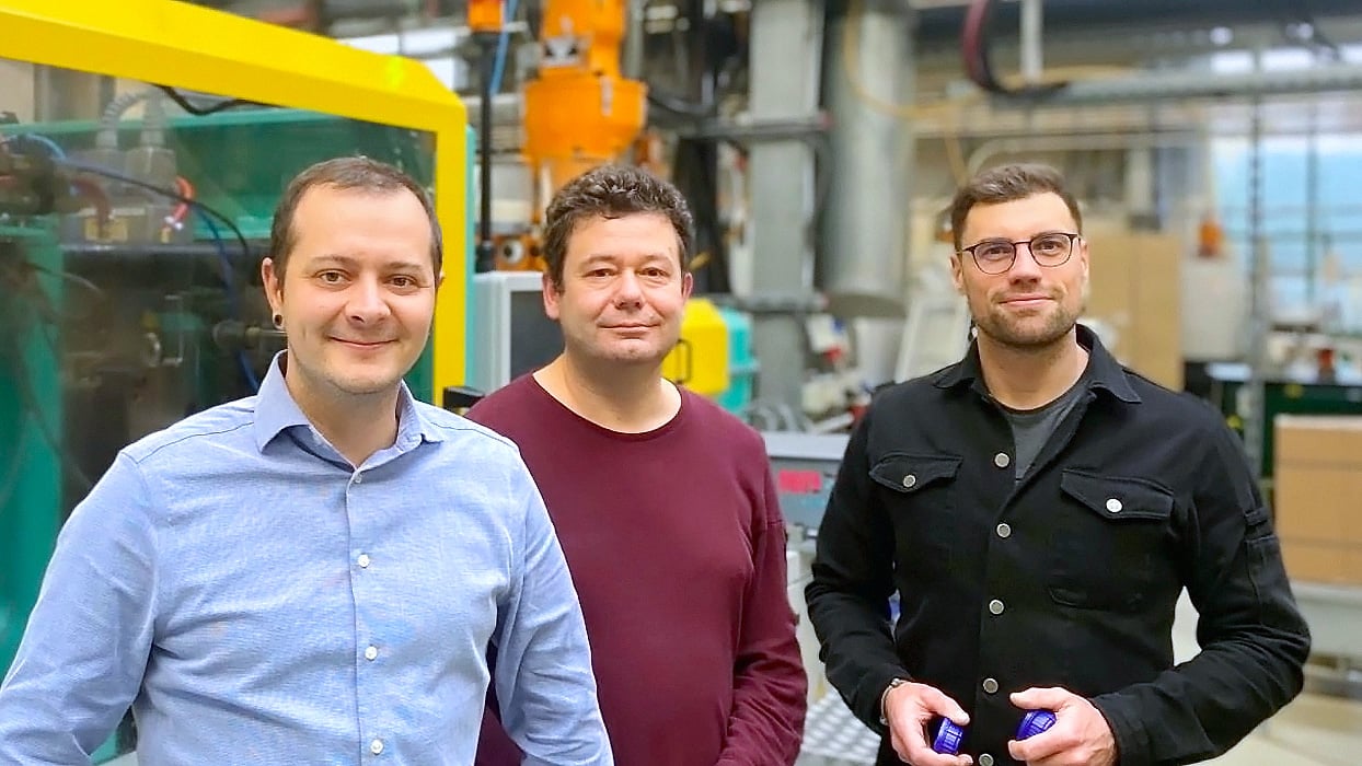 Three men stand together in a plastics manufacturing facility. They smile at the camera, with injection molding machines and industrial equipment visible in the background. One holds a small blue plastic cap in his hands.