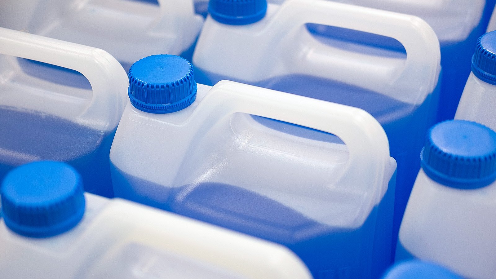 Close-up of white plastic jerrycans used as industrial packaging, filled with blue liquid and sealed with blue screw caps, arranged in rows for storage and transport.
