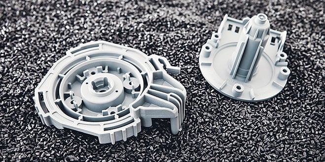 Two light grey injection-moulded plastic parts with detailed geometry lie on a bed of dark plastic granules, illustrating lightweight component manufacturing.