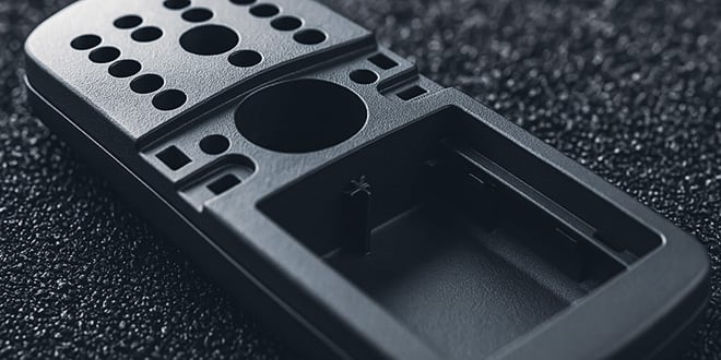 Close-up of a dark grey plastic electronic housing with precision-cut holes and recesses, placed on a surface of plastic granules, illustrating high-quality injection moulding.