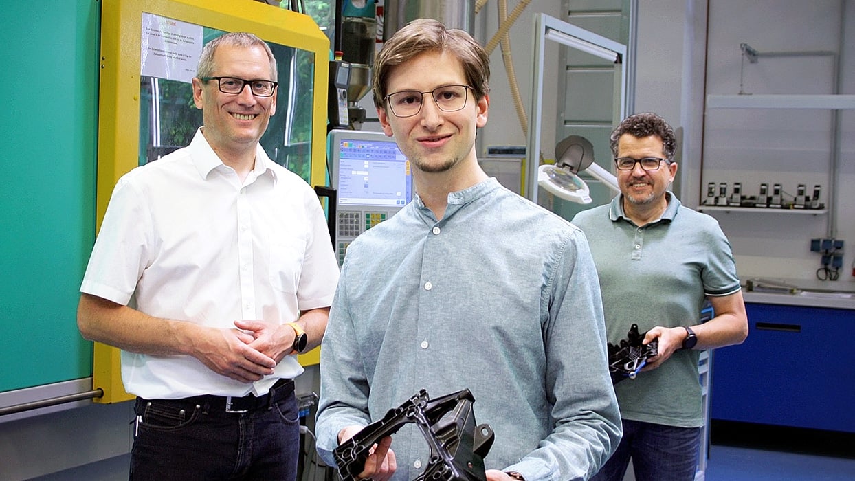 Three men stand in a manufacturing workshop, smiling at the camera. Two hold black plastic parts while machinery and equipment are visible in the background.