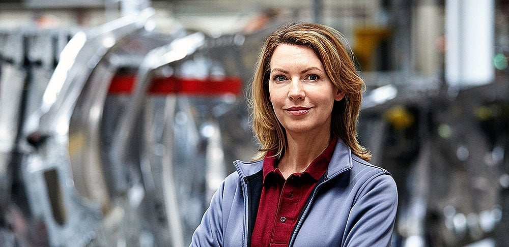 A confident woman in a gray jacket and red shirt stands in front of large industrial machinery, looking directly at the camera with a slight smile.