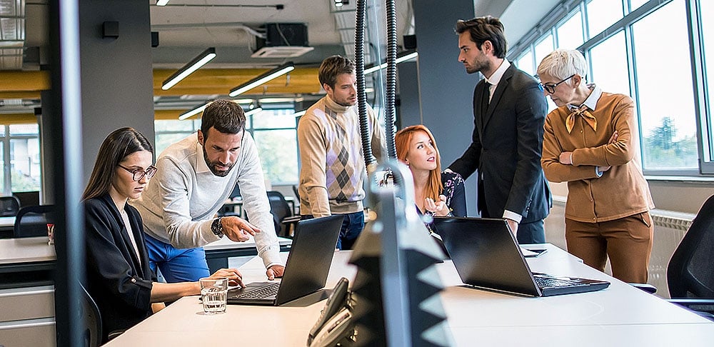 A diverse group of six colleagues gathers around laptops in a modern office, engaged in a collaborative discussion.