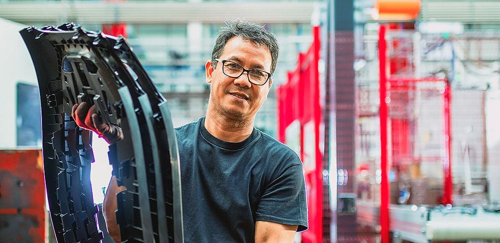 A man wearing glasses and a black shirt holds a large plastic automotive part inside a brightly lit factory.