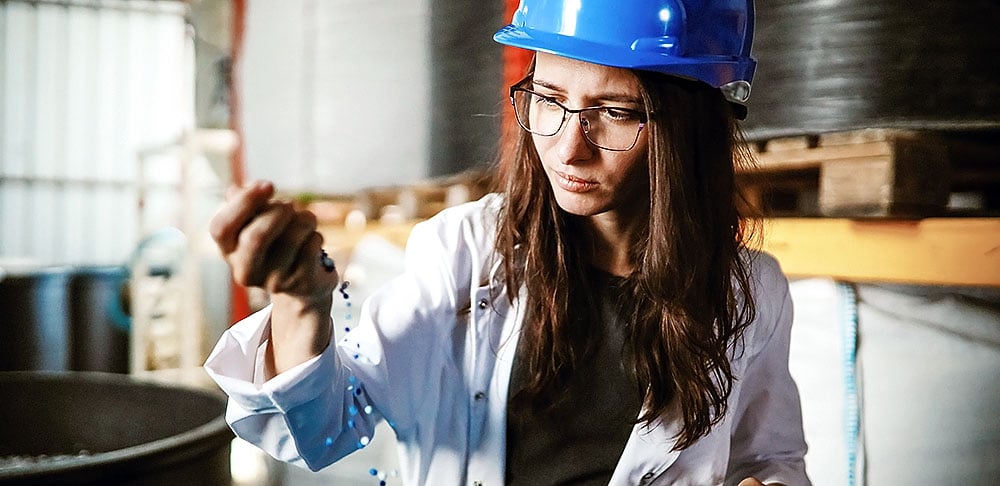 A woman wearing glasses, a blue hard hat, and a white lab coat examines plastic pellets in her hand at an industrial site.